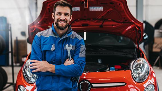 Un garagiste en bleu de travail devant une voitiure rouge au capot relevé tient une clef anglaise